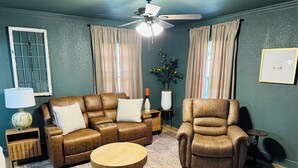 A living room with a brown leather sofa, a wooden coffee table, and a leather armchair. There is a ceiling fan, a lamp, and a vase with flowers on a side table.