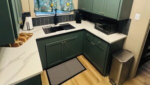 A compact kitchen with dark green cabinets, a white countertop, a patterned window curtain, and a stainless steel trash bin.