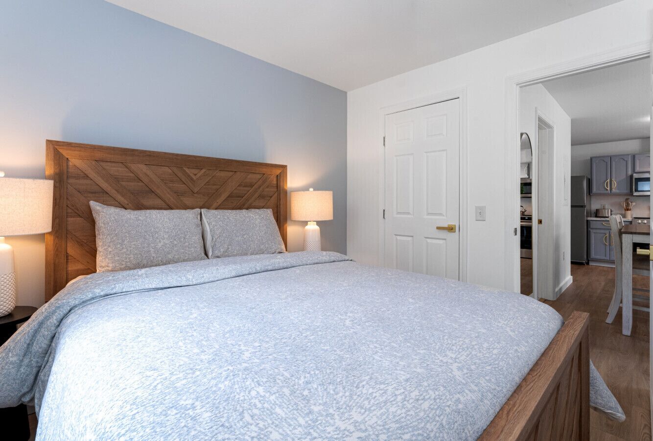 A bedroom with a wooden headboard, a bed with a patterned bedspread, two bedside lamps, and a white door leading to a kitchen area.
