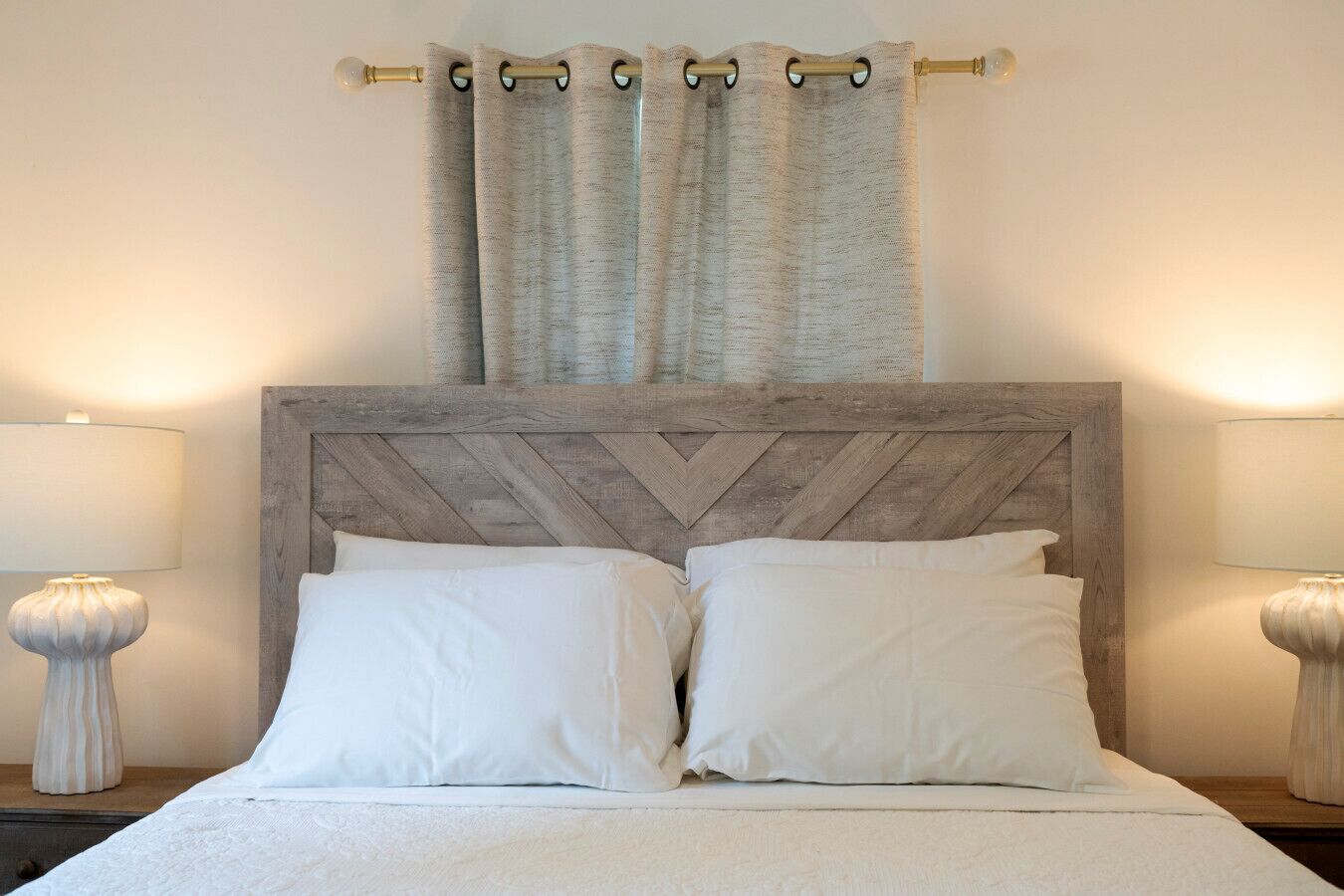 Una cama bien hecha con cabecera de madera, dos lámparas de noche y una cortina.