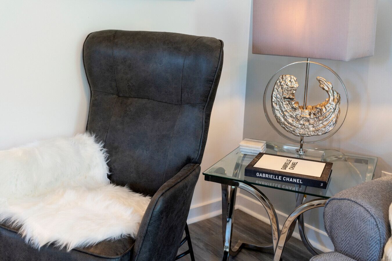 Un sillón de cuero negro con un cubrepiedal de piel blanca, una mesita de vidrio con un libro titulado 'Gabrielle Chanel', y una escultura decorativa de metal.