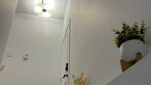 Un pasillo con una lámpara en el techo, una puerta, un equipo de aire acondicionado pequeño y una planta en maceta sobre un estante.