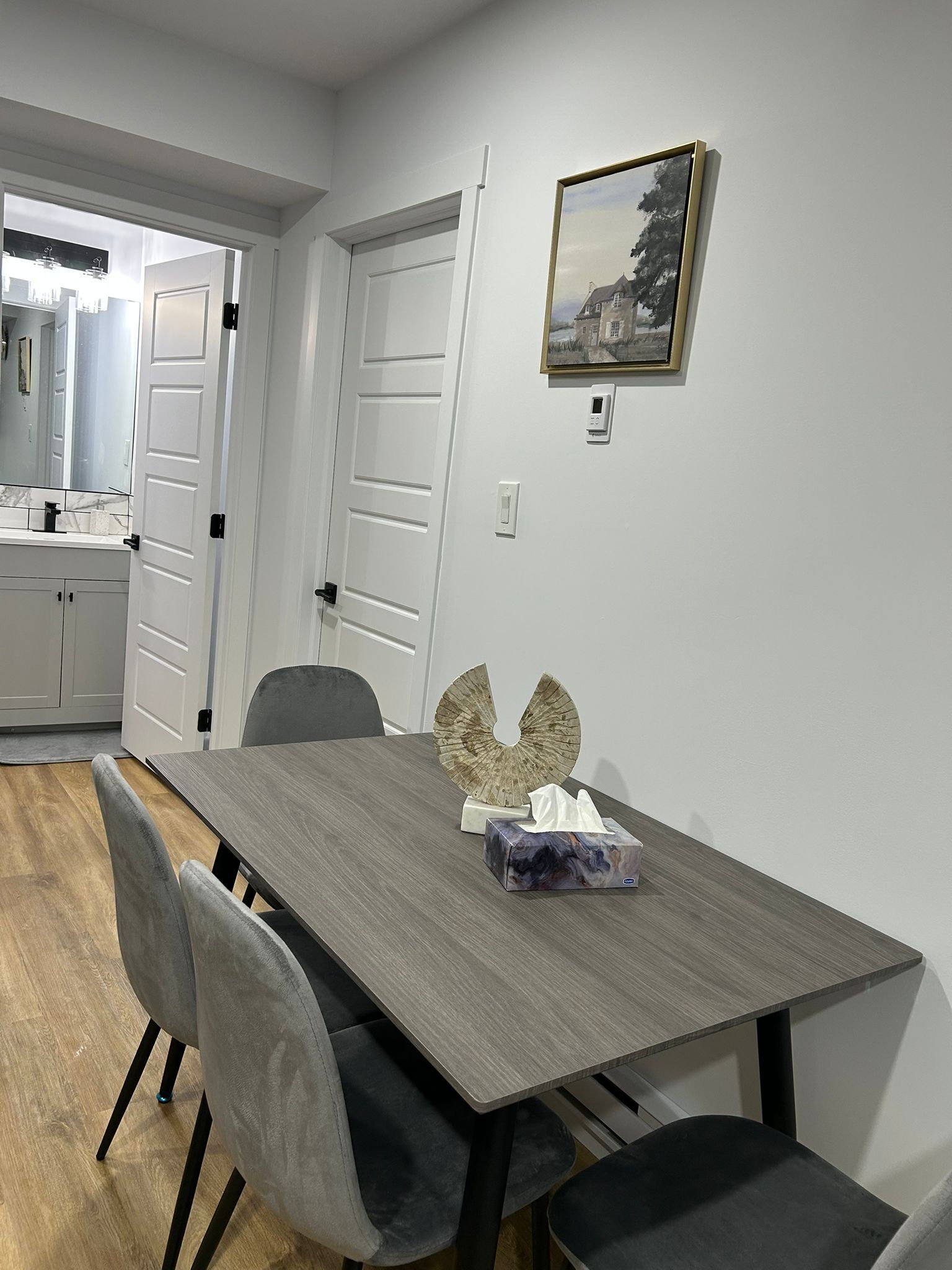 Un coin repas comprenant une table en bois, des chaises à revêtement gris, un élément décoratif mural et un tableau.