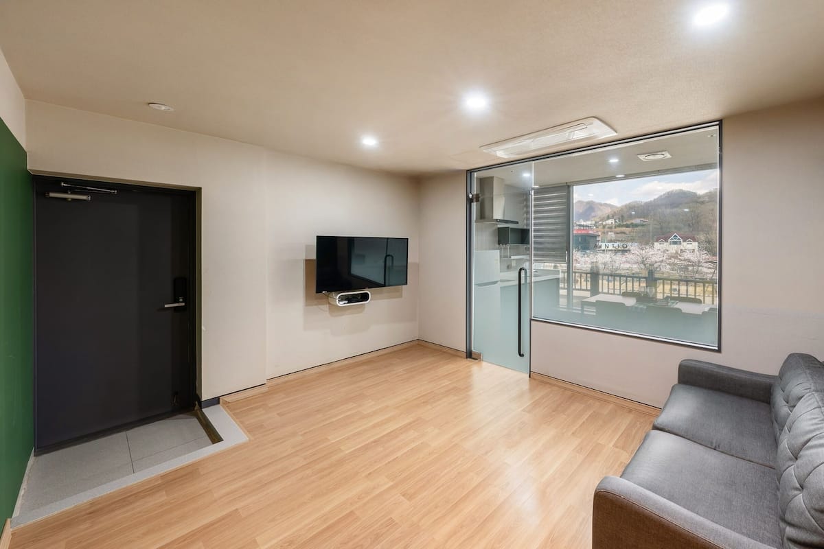 Una habitación moderna con suelo de madera, un sofá gris, un televisor montado en la pared y un amplio ventanal con vistas a un pueblo.