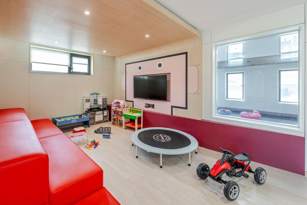 Una sala de estar moderna con un sofá rojo, una zona de juegos con juguetes y un trampolín, y un televisor grande montado en la pared.