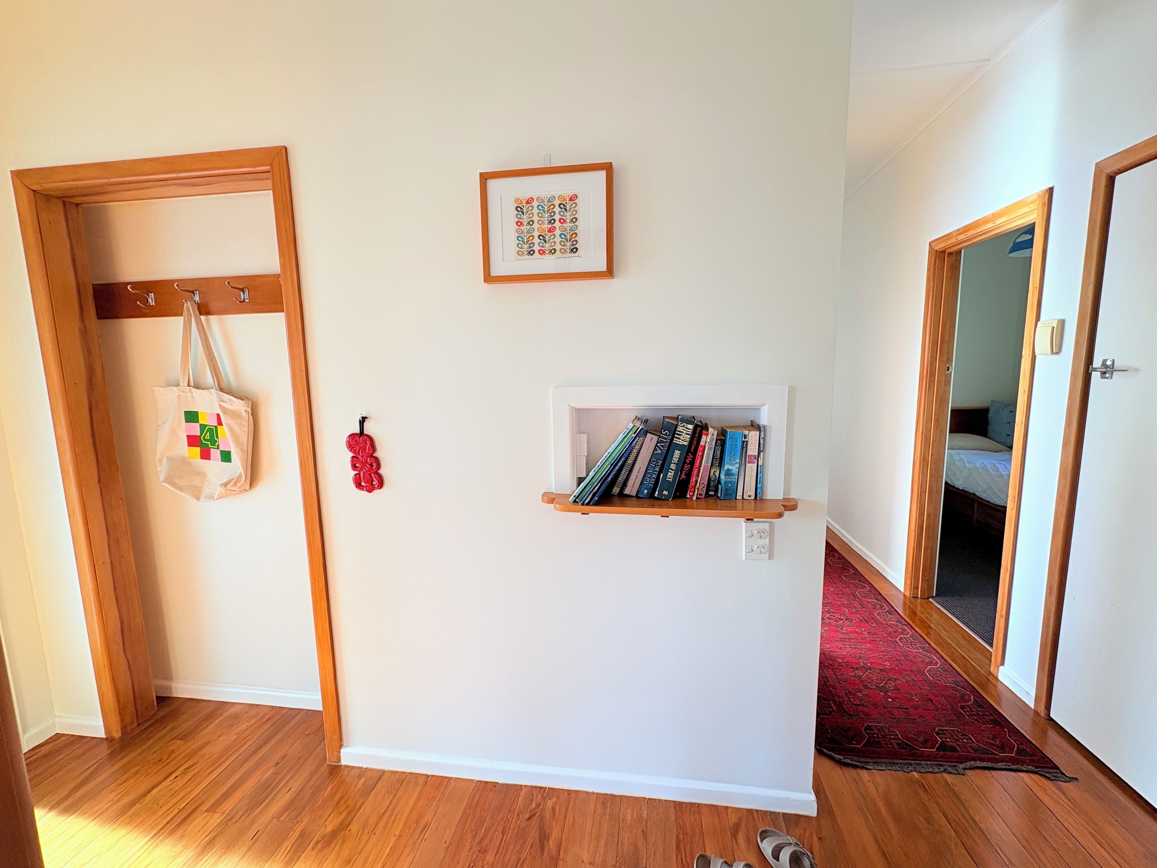 Un corridoio con pavimento in legno, parete bianca, una piccola libreria con libri, un quadro incorniciato, una borsa appesa e una porta che dà su un’altra stanza.