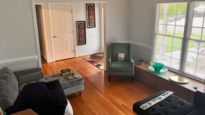 Un salon doté d’un parquet, d’un canapé gris, d’un fauteuil vert et d’un pouf noir. On y trouve une fenêtre avec des stores, une porte et des œuvres d’art décoratives accrochées au mur.