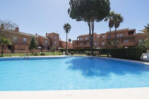 A swimming pool surrounded by apartment buildings with balconies, palm trees, and a clear sky.