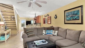 A living room with a sectional sofa, a coffee table, a ceiling fan, a staircase, and a kitchen in the background.