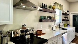 A modern kitchen with a stainless steel stove, oven, and a built-in range hood. There are wooden shelves above the counter displaying various kitchen items and plants.
