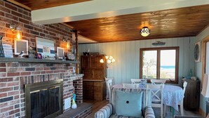 A cozy living room with a brick fireplace, wooden ceiling, and a dining area with a view of the beach. - Oceanfront Escape: Deck & Granite Steps to Shore (Harpswell)