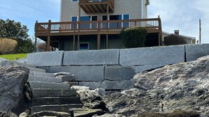 A two-story house with a wooden deck and a stone retaining wall. - Oceanfront Escape: Deck & Granite Steps to Shore (Harpswell)