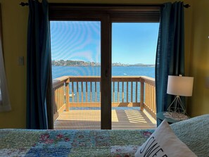 A bedroom with a large window overlooking a wooden deck and a body of water. - Oceanfront Escape: Deck & Granite Steps to Shore (Harpswell)