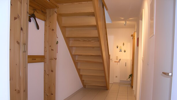 Una escalera de madera que sube a un segundo piso, un pasillo con baldosas y una puerta con una planta pequeña.