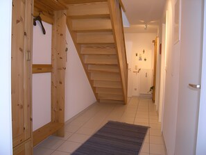 Een houten trap die leidt naar een tweede verdieping, een betegelde gang en een deur met een klein plantje.