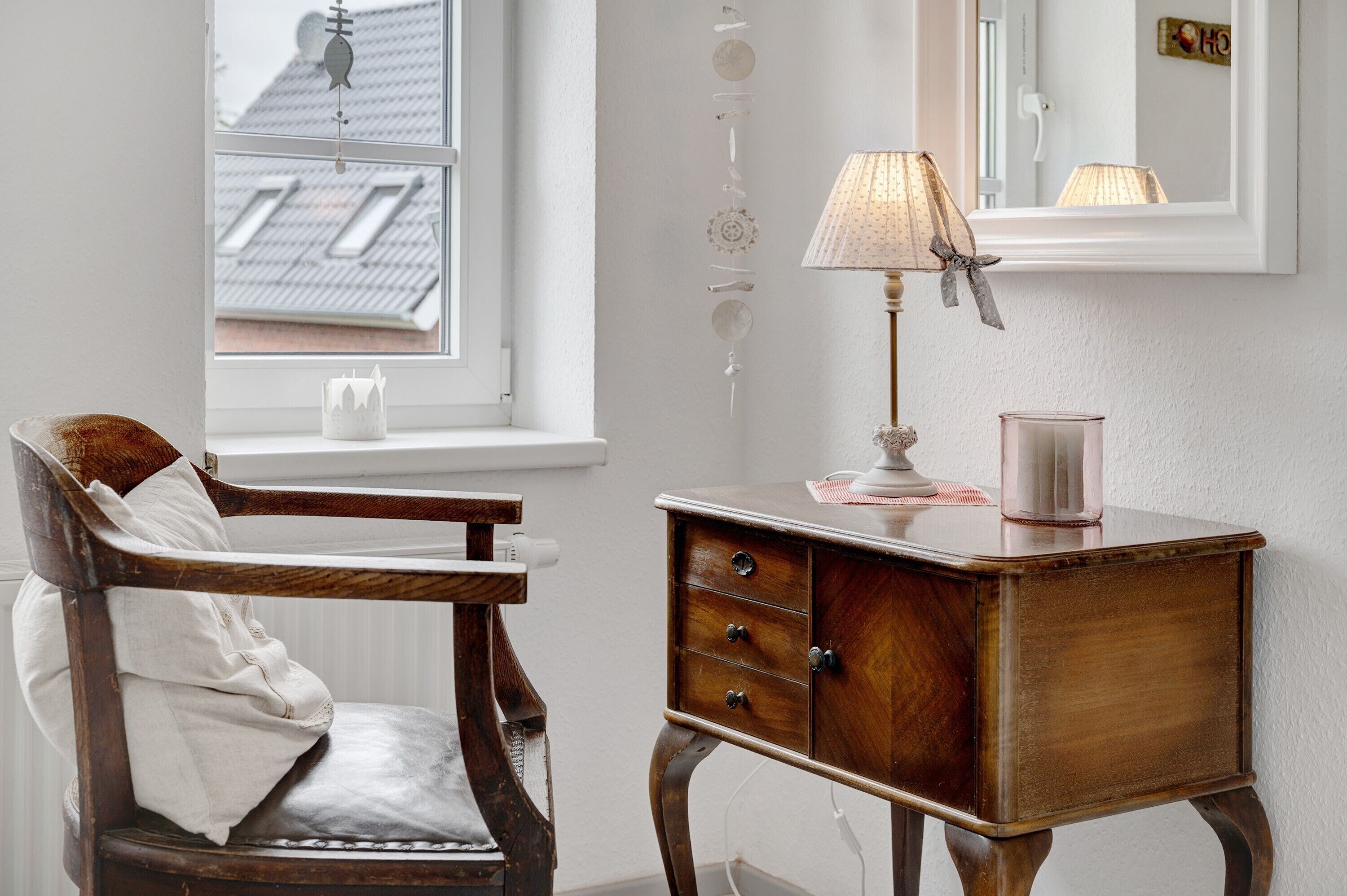 Une chaise en bois avec un coussin, une petite armoire en bois avec des tiroirs, une lampe et une fenêtre donnant sur un immeuble.