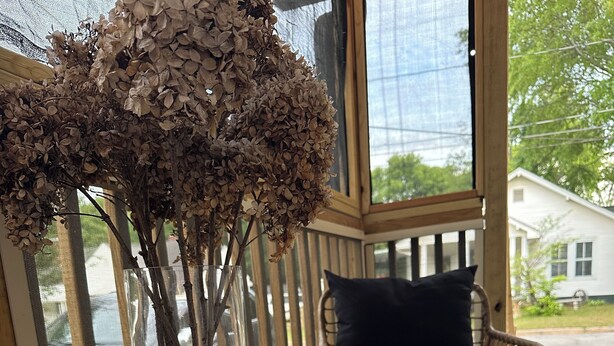 Une table en verre sur laquelle reposent deux verres à vin et un vase de fleurs séchées, le tout placé sur une véranda avec une chaise en osier et un coussin noir.