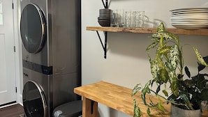 Une buanderie équipée d’une machine à laver et d’un sèche-linge superposés, d’étagères en bois où sont rangés des verres, et d’une plante en pot.
