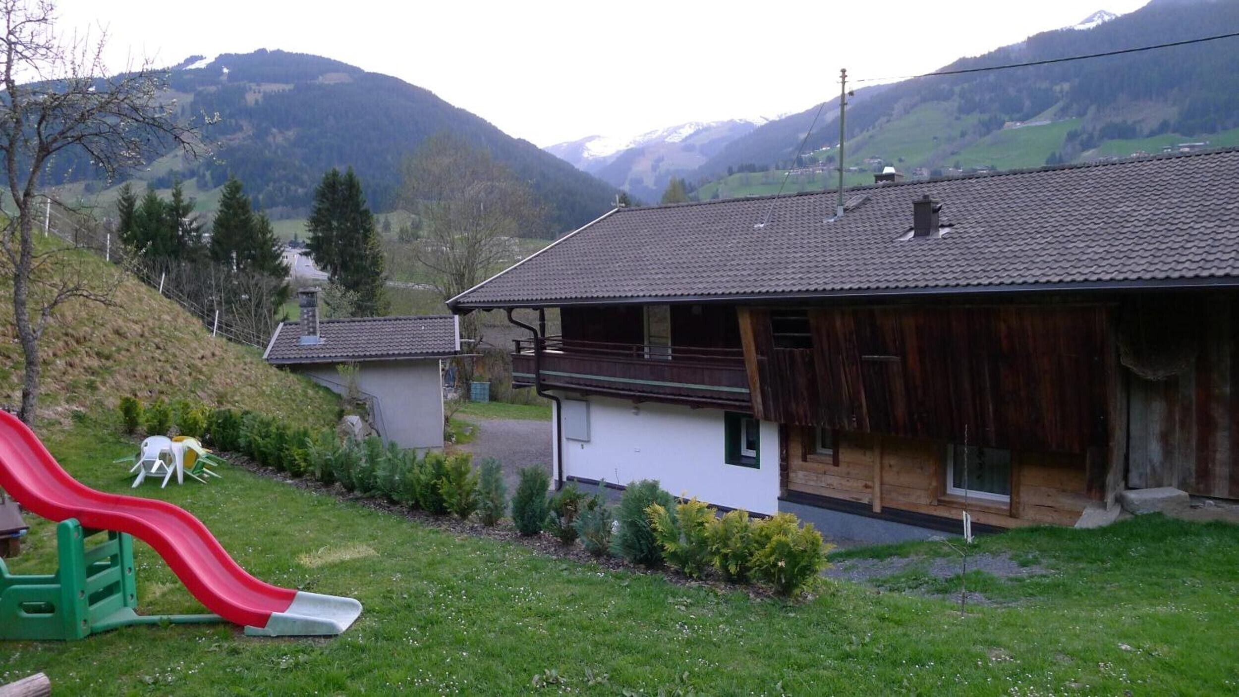 A house with a red slide in the yard, surrounded by greenery and mountains.