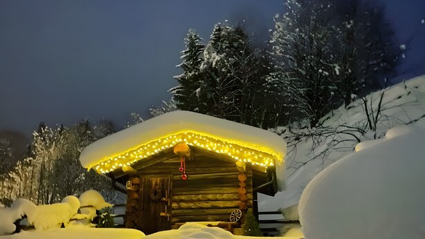 A snow-covered cabin with a roof adorned with lights, surrounded by snow.