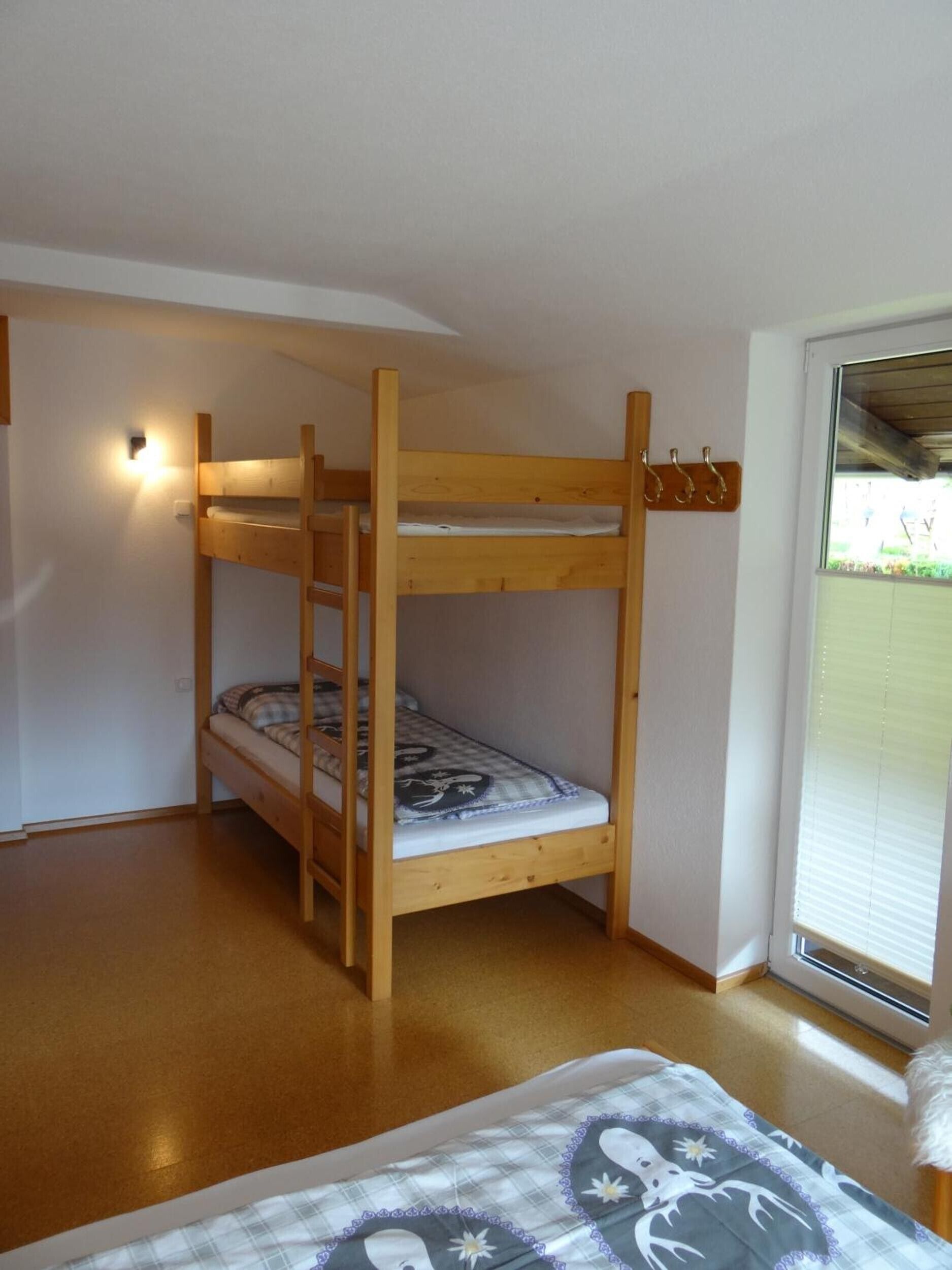 Un lit superposé avec une couverture à carreaux, une lampe fixée au mur et une fenêtre avec des stores.