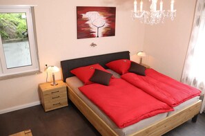 A bedroom with a bed, two bedside tables, a nightstand, a chandelier, a painting, and a window.