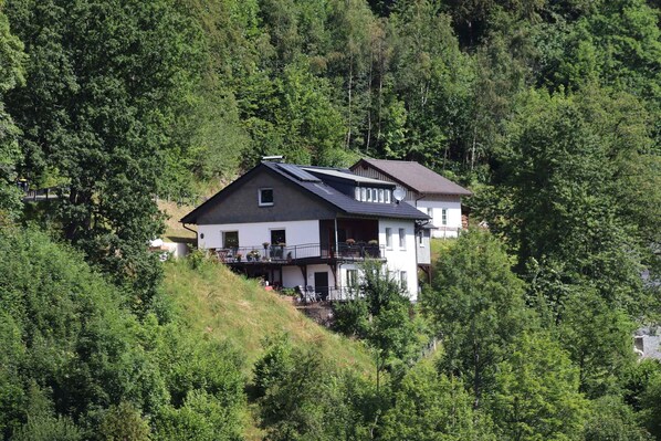 A two-story house with a balcony, surrounded by dense forest.