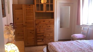 Ein Schlafzimmer mit einem hölzernen Kleiderschrank, einem Bett mit gemusterten Betttextilien, einem Nachttisch, einer Lampe und einem Fenster mit Vorhängen.
