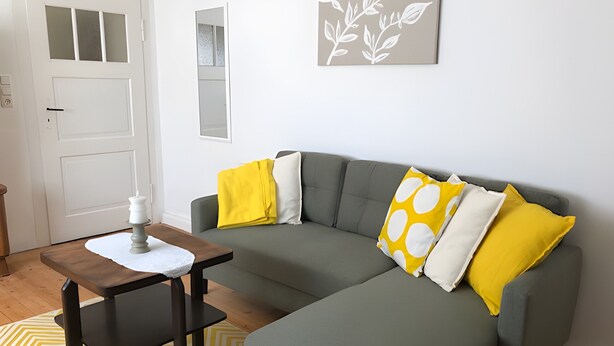 Ein Wohnzimmer mit einem grauen Sofa, einem kleinen Tisch mit einer Kerze und einem gelb gemusterten Teppich.