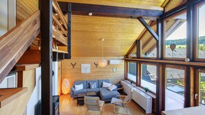 A modern interior with a wooden staircase, a sectional sofa, a dining table, and large windows offering a view of the outdoors.