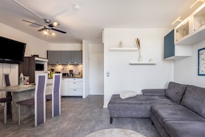 Un salon moderne avec un canapé gris, un coin repas avec une table et des chaises, une cuisine avec des meubles gris et un ventilateur de plafond.