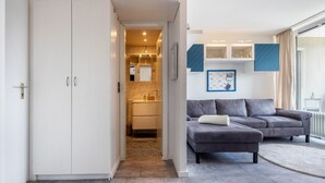 Una sala de estar moderna con un sofá gris, un armario blanco y una cocina visible a través de una puerta abierta.