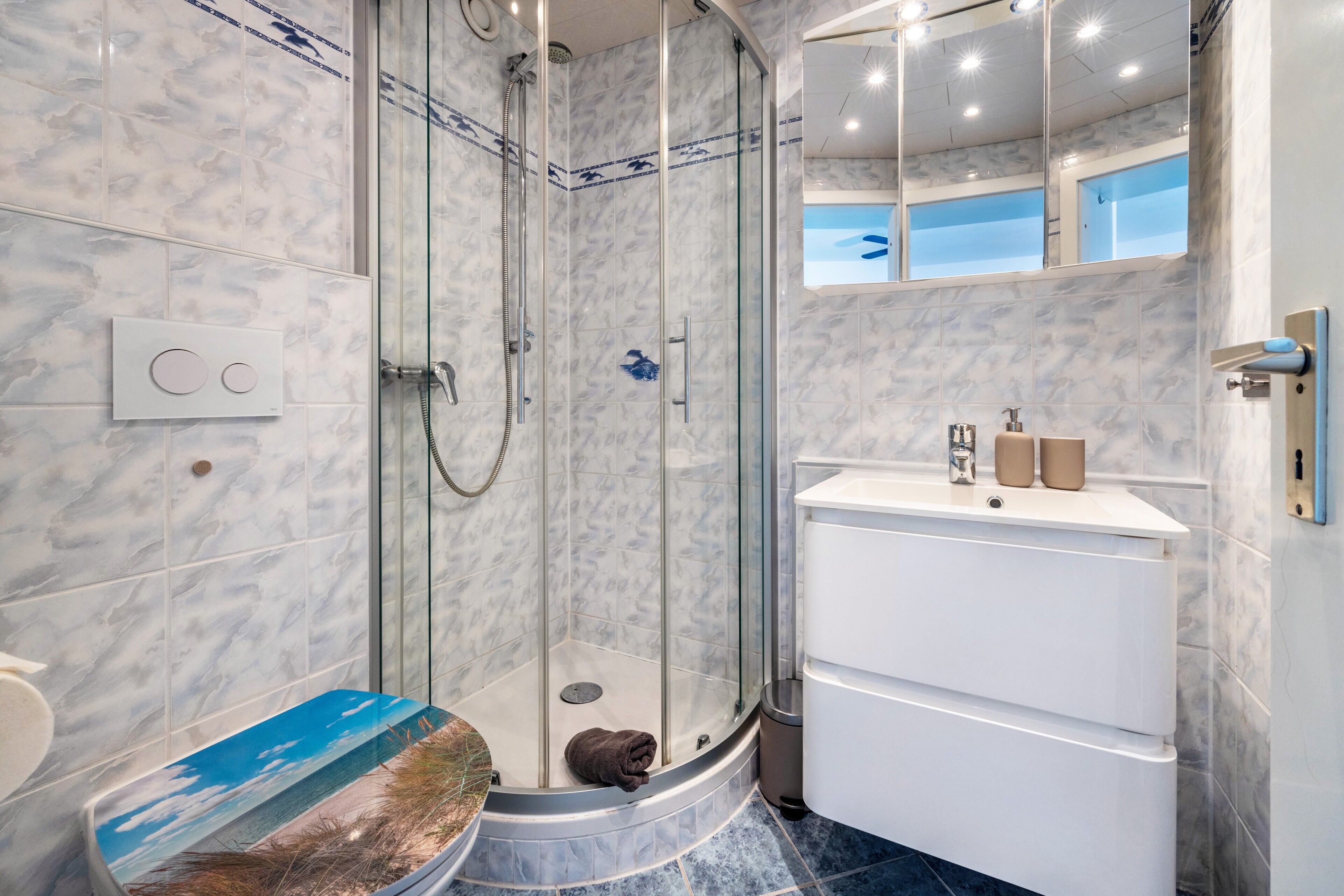 Un baño moderno con ducha envuelta en vidrio, un lavamanos blanco con dos botellas y una pared con azulejos de mármol.
