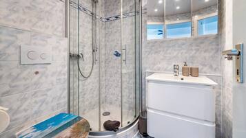 Un baño moderno con ducha envuelta en vidrio, un lavamanos blanco con dos botellas y una pared con azulejos de mármol.