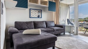Una sala de estar moderna con un sofá gris oscuro, un cojín blanco, un cuadro en la pared y vistas al exterior a través de un ventanal.