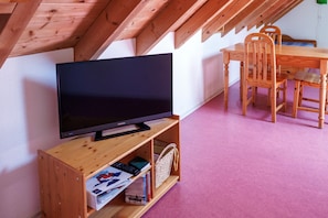 Huoneessa on televisio, joka on sijoitettu puiseen jalustaan, ruokapöytä tuoleineen sekä vaaleanpunainen, matolla peitetty lattia. Huoneen katossa on näkyvillä puupalkit.