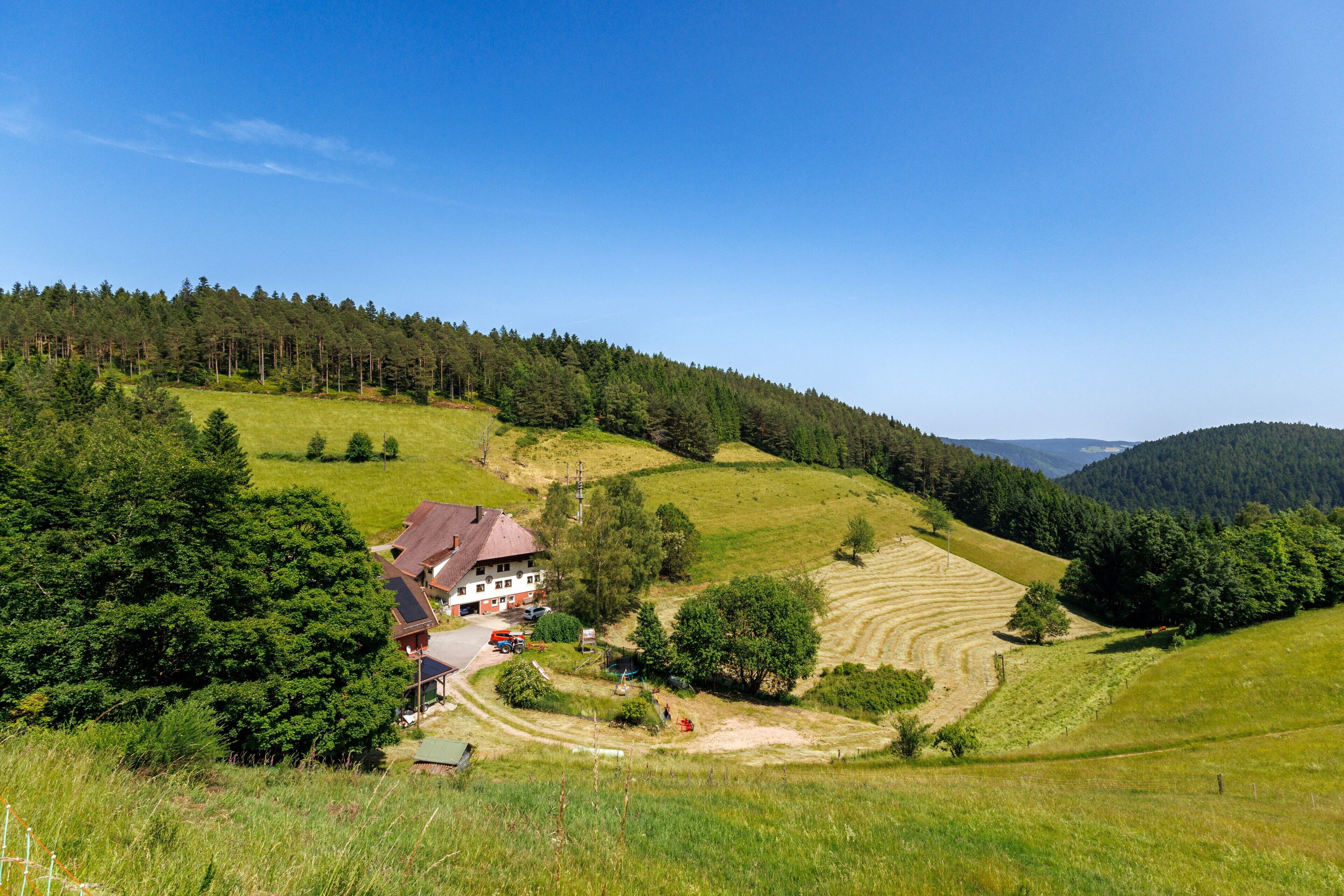Ein ländliches Haus mit rotem Dach, umgeben von grünen Feldern und einem bewaldeten Hügel.