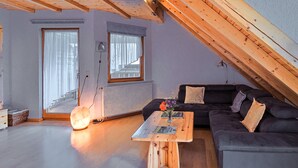Ein Loft-Zimmer mit Holzbalkendecke, einem dunkelgrauen Sofa, einem Holztisch und einem großen Fenster mit Vorhängen.