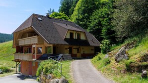 Ein zweistöckiges Holzhaus mit Balkon, befestigter Auffahrt und bewaldeter Hanglage.