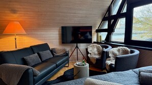 Een moderne woonkamer met een zwarte leren bank, een grijze fauteuil, een klein rond tafeltje met een fles en een aan de muur bevestigde televisie.