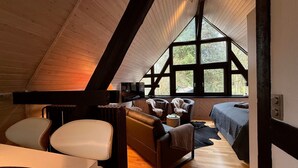 Een gezellig slaapkamer op zolder met een groot raam, houten balken, een bed, een bank en een tafel.