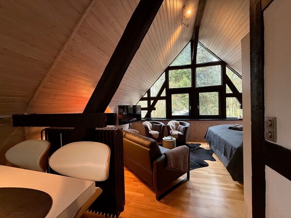 Un acogedor dormitorio en el ático con un ventanal grande, vigas de madera, una cama, un sofá y una mesa.