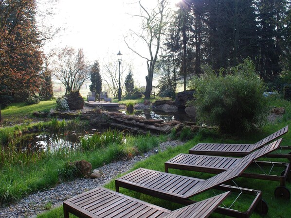 A serene outdoor area with a pond, wooden lounge chairs, and a stone pathway.