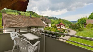 A balcony with a table and chairs, overlooking a green valley with houses and a winding road. - Gästehaus Gerda: Double Room 4 with Mountain View and Wi-Fi (Bad Peterstal-Griesbach)