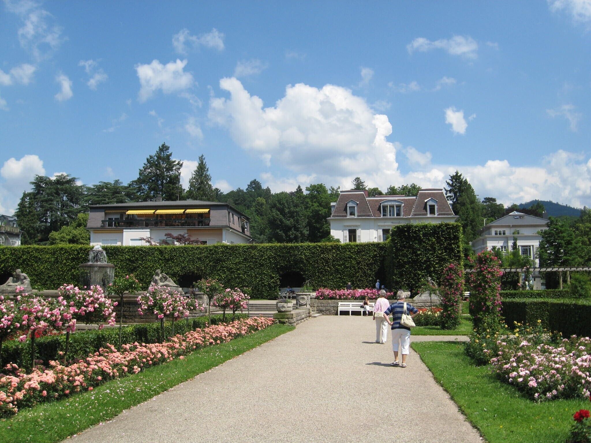Un chemin menant à un bâtiment doté d’un jardin où poussent des fleurs roses et bordé d’une haie.