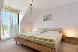 Una camera da letto con un letto, comodini, un termosifone, una finestra con tende leggere e una pianta pensile.
