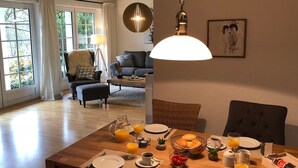 Zona pranzo con tavolo di legno apparecchiato per la colazione, un divano e una lampada sullo sfondo.