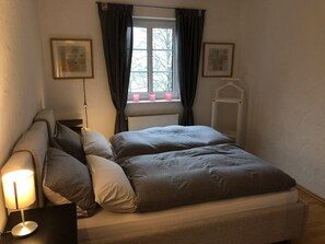 Ein Schlafzimmer mit einem Bett, einer Nachttischlampe, einem Fenster mit Vorhängen und zwei gerahmten Bildern an der Wand.