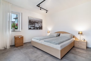 A bedroom with a bed, bedside lamps, a nightstand, a wicker basket, a window with curtains, and a cityscape picture on the wall.