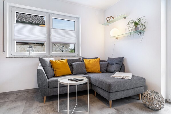 A modern living room with a grey sectional sofa, a small white side table, a round coffee table, a potted plant, and a decorative wall shelf.